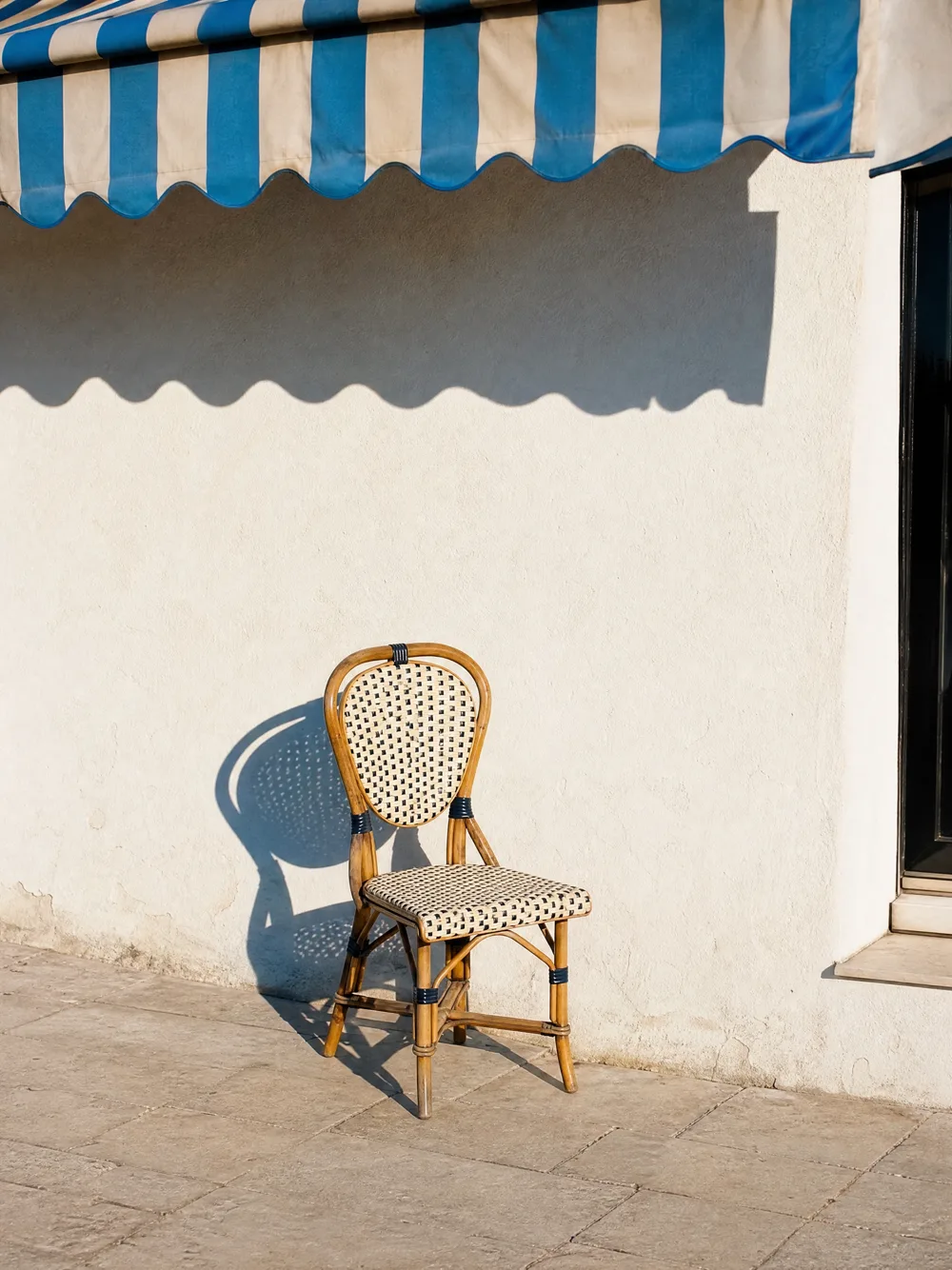 Une chaise en rotin au soleil devant un mur blanc, son ombre bleue au sol